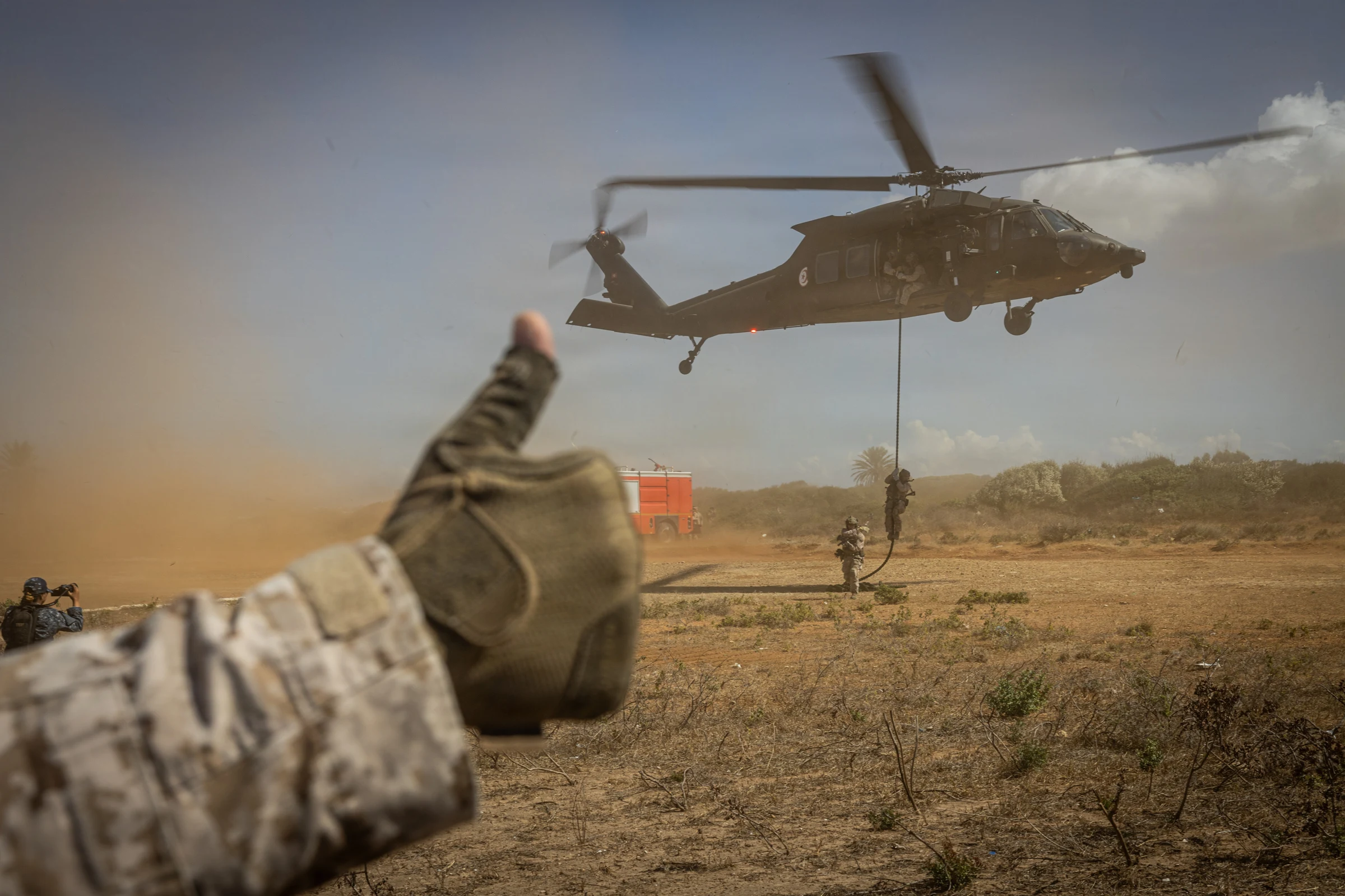 Service members slide down ropes from a hovering UH-60 helicopter toward a paved surface during maritime boarding training.