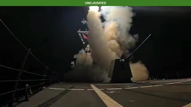 A missile rises in a plume of smoke beside a deck gun on a darkened warship at night.