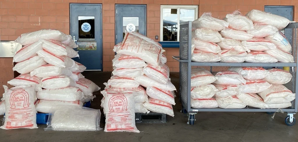 Stacks of white woven sacks printed with red lettering, piled on pallets and a metal cart in front of a brick wall with blue doors at a port inspection area.