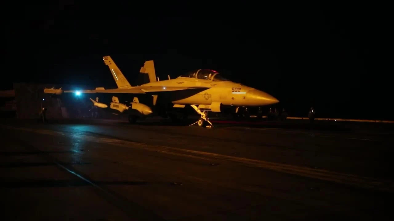 Night view of a grey U.S. Navy F/A-18 fighter jet on an aircraft carrier deck, lit by deck lights against a black sky, moments before launch.