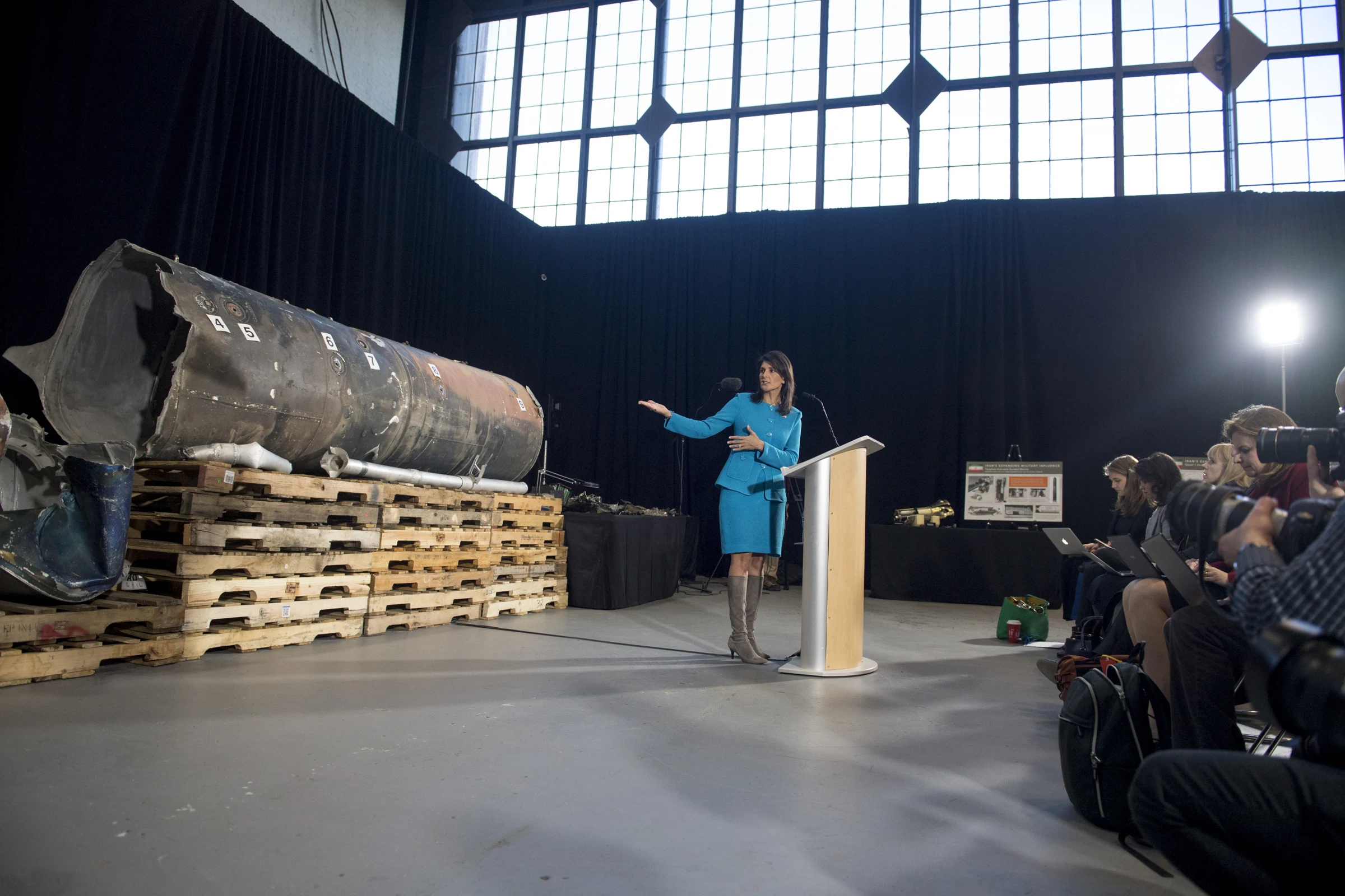 A woman in a blue suit gestures toward a large, scorched cylindrical missile section resting on wooden pallets in a hangar, while photographers and reporters sit at right.