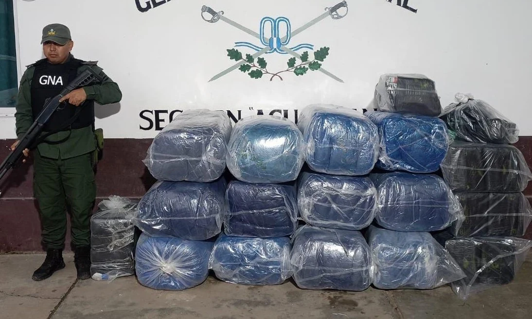 Argentine gendarme in green uniform and GNA vest stands beside stacked plastic-wrapped coca-leaf bundles indoors under a Gendarmería wall seal.