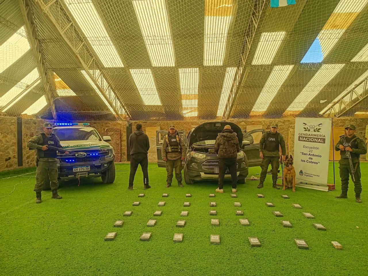 Argentine gendarmes stand behind rows of seized cocaine packages laid on a green floor, with a patrol truck, the opened SUV and a police dog; two detainees face away from the camera.