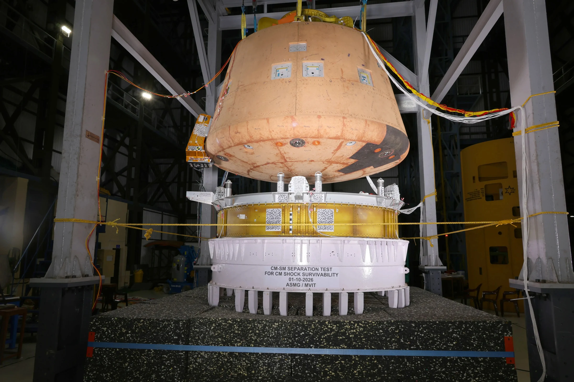Orange conical Gaganyaan crew module suspended above a white cylindrical service-module stand labeled CM-SM SEPARATION TEST inside a test hall.