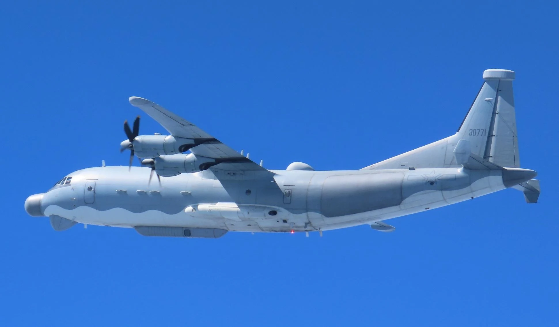 A grey four-engine turboprop military aircraft with a long nose radome and antennas on its fuselage flies left across a clear blue sky.