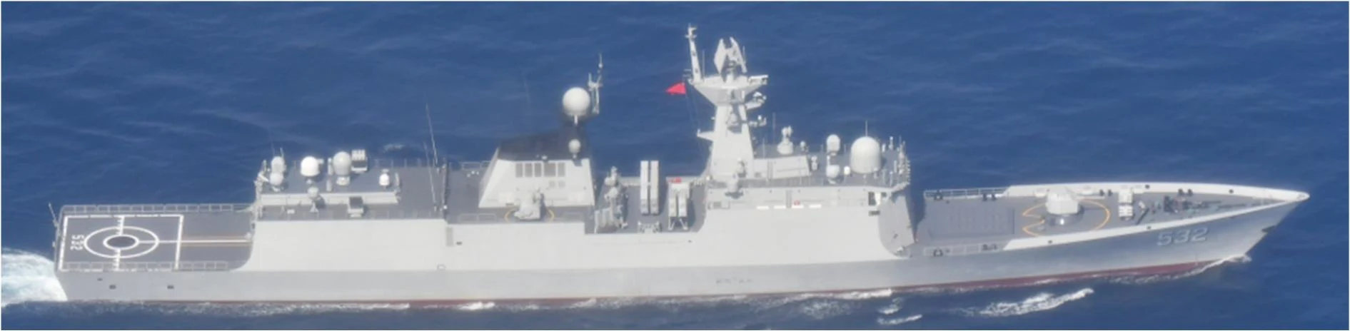 Grey Chinese navy frigate in side view on deep blue sea, with hull number 532 on the bow, a helicopter deck marked with a circle at the stern, radar domes and a mast flying a red Chinese flag, and white wake at the stern.