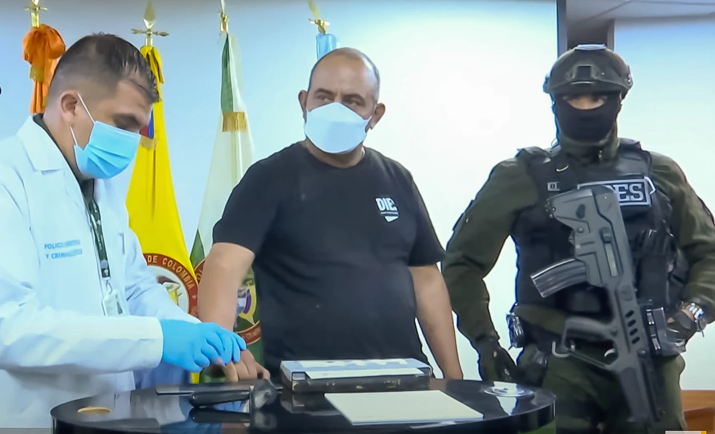 A man in a black T-shirt and face mask stands at a table while a forensic officer in a lab coat takes his fingerprints, beside a masked, helmeted officer holding a rifle, with Colombian flags behind.