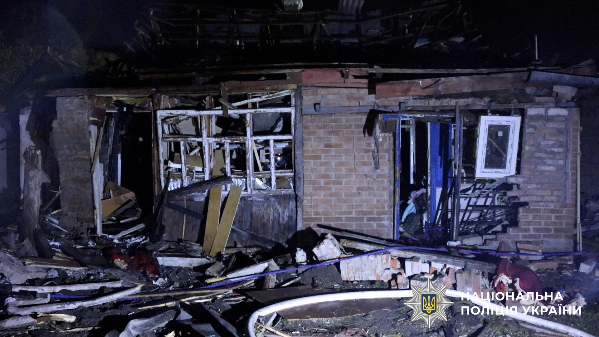 A single-storey brick house at night with its windows and roof blown out and debris piled in the yard, National Police of Ukraine watermark at lower right.