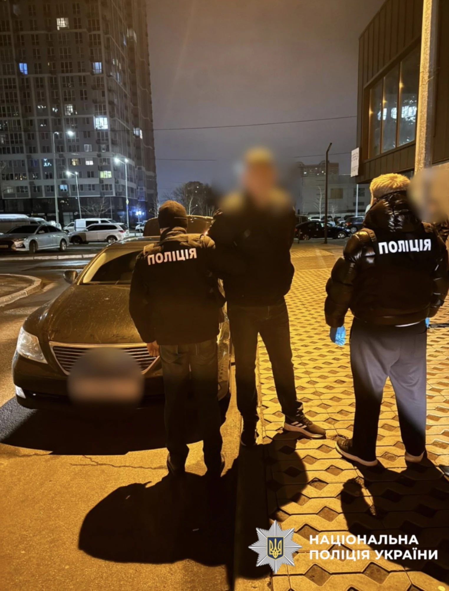Night street photo: two police officers in black jackets marked 'ПОЛІЦІЯ' stand beside a man in a dark jacket whose face is blurred, next to a dark sedan with a blurred number plate, with lit high-rise apartment blocks behind them and a National Police of Ukraine watermark.
