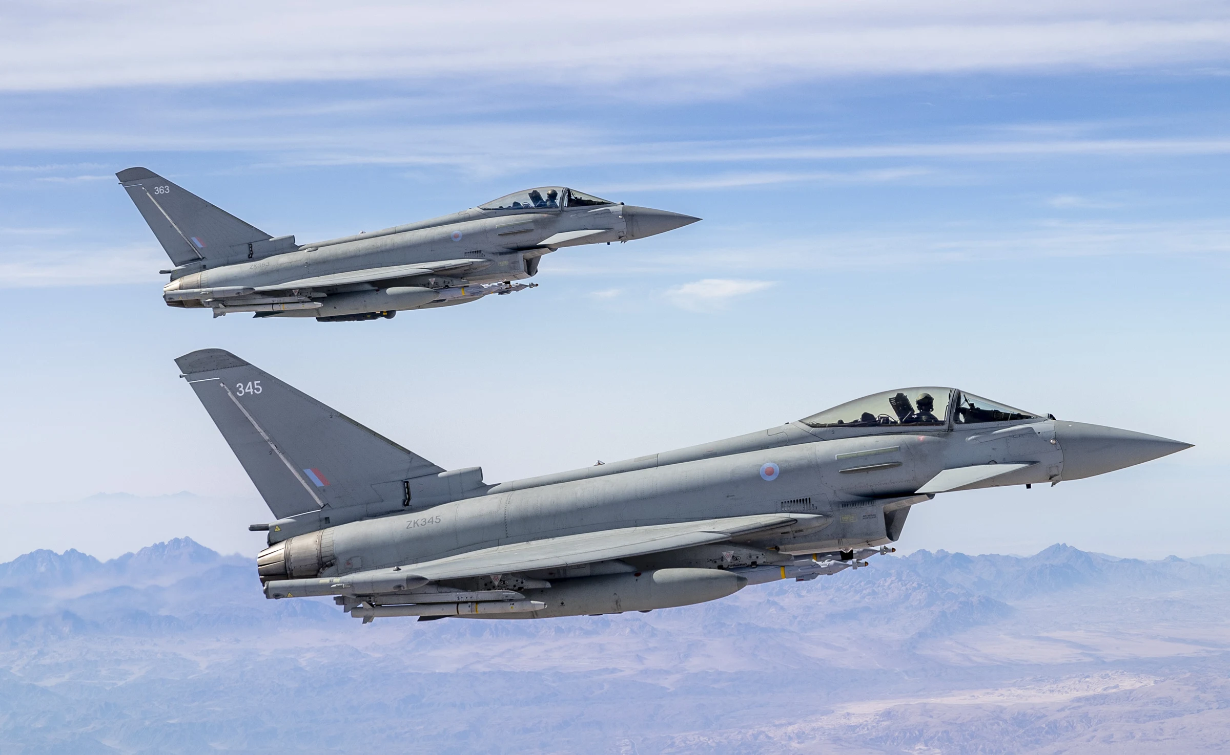 Two grey RAF Typhoon fighter jets flying in formation above a hazy desert mountain range under a pale blue sky, one slightly above and behind the other, with the nearer jet's tail number 345 visible.
