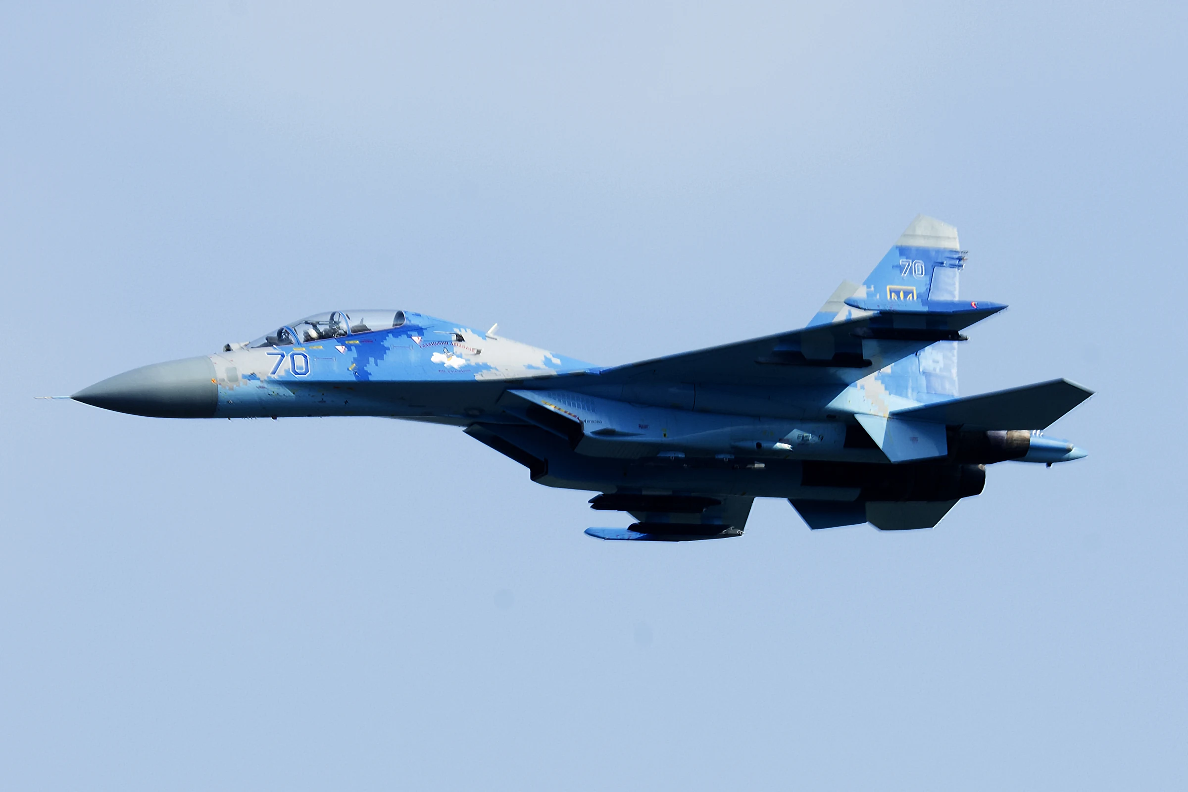 A two-seat fighter jet in blue and grey digital camouflage with the side number 70 flies left across a pale blue sky.