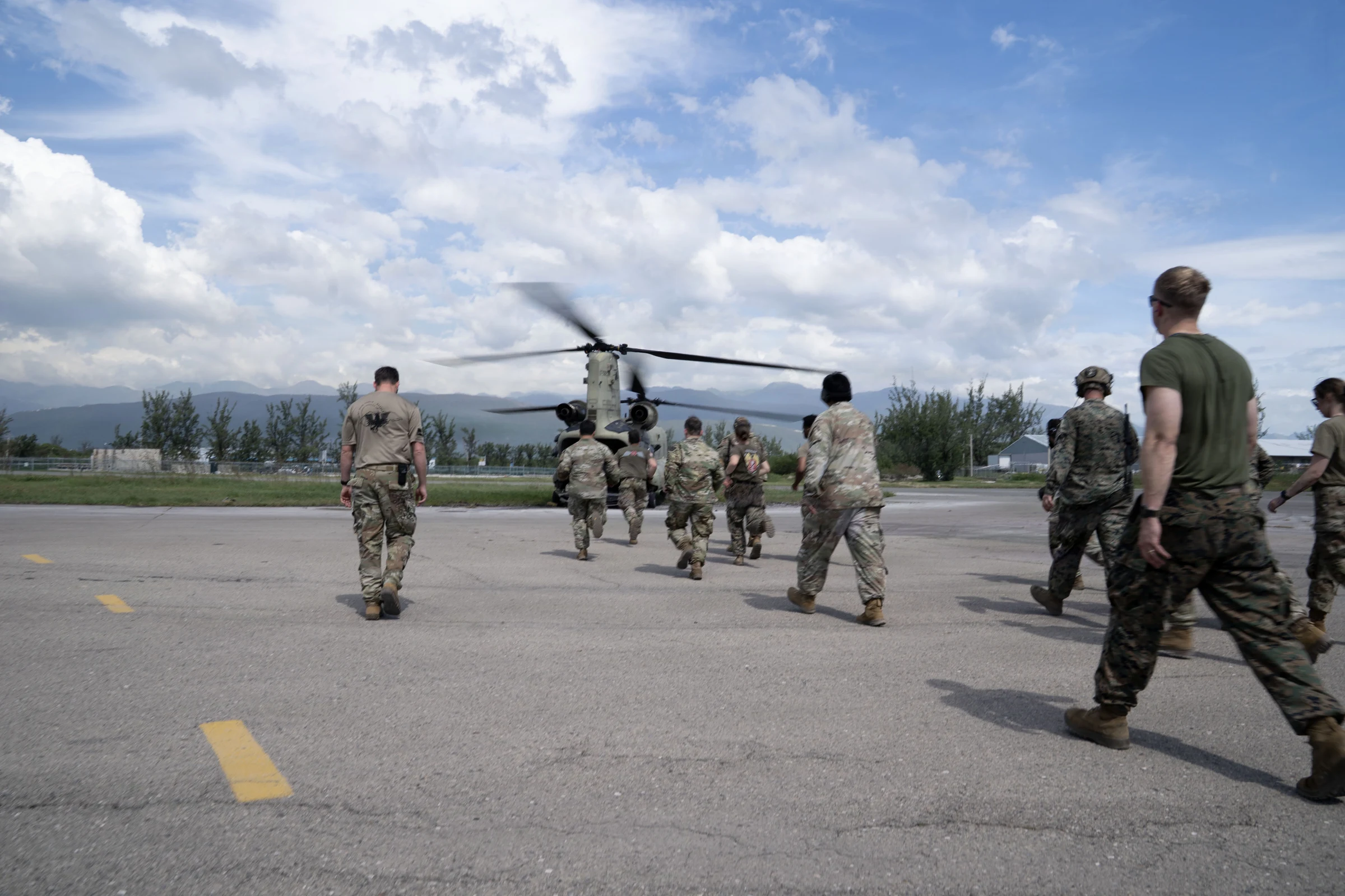 U.S. service members in camouflage uniforms walk across an airport apron in a line toward a CH-47 Chinook helicopter with its rotors turning, under a blue sky with white clouds and green hills in the distance.