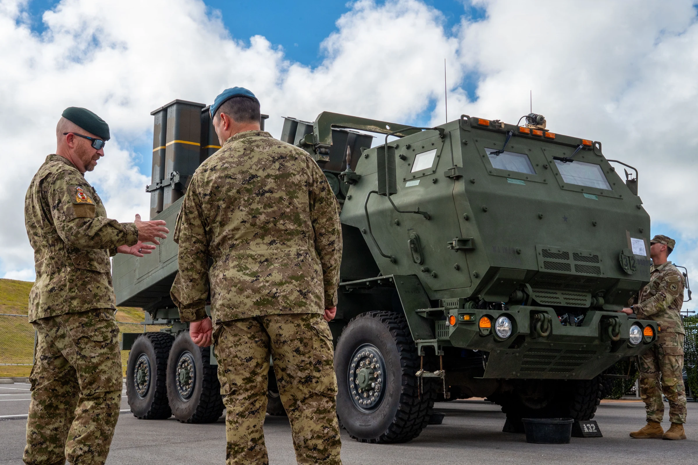 Two soldiers in Canadian camouflage and green berets talk beside a green U.S. Army HIMARS rocket-launcher truck parked on a road, with a third service member nearby and white clouds over green hills behind.