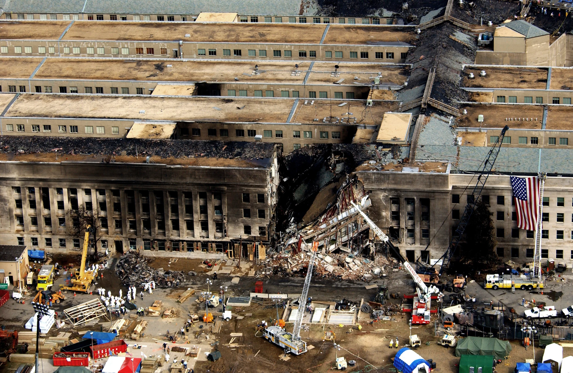 Aerial photo of the Pentagon's damaged facade: a collapsed, blackened section with exposed floors and rubble, cranes and a fire-truck ladder reaching in, a large U.S. flag hanging on the wall to the right, and rescue workers, tents and vehicles on the ground in front.
