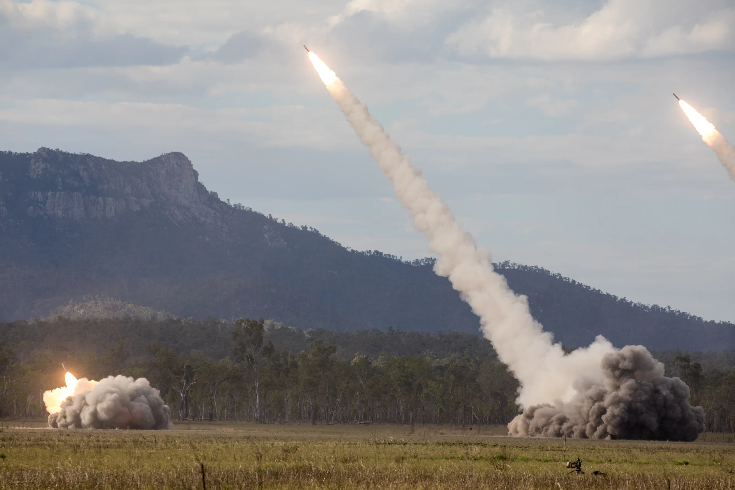 Rockets streak upward on long white smoke trails from launchers hidden in dust clouds on a grassy plain, with a rocky forested ridge and cloudy sky behind and a third launch flashing at the left.