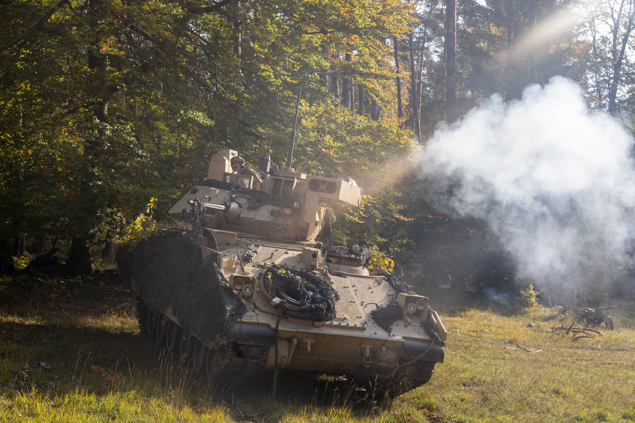 A tan U.S. Army M2 Bradley fighting vehicle draped in camouflage netting sits at the edge of a forest clearing with a crew member in the turret, as white smoke billows to its right and sunlight filters through autumn trees.