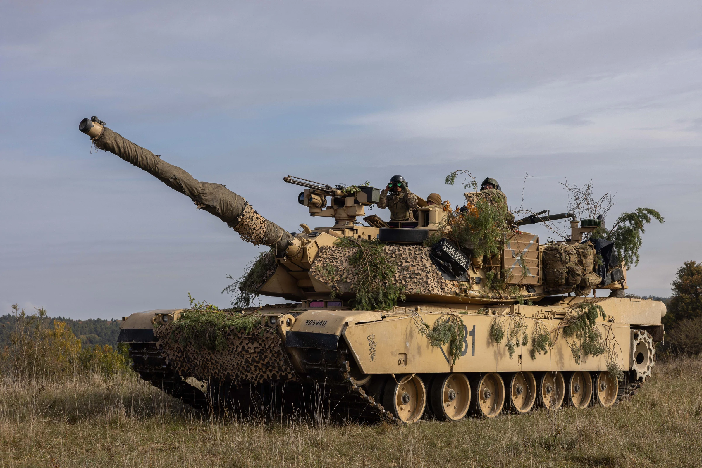 U.S. M1 Abrams tank covered in camouflage netting and foliage in a grass field, two soldiers in the turret, one using binoculars.