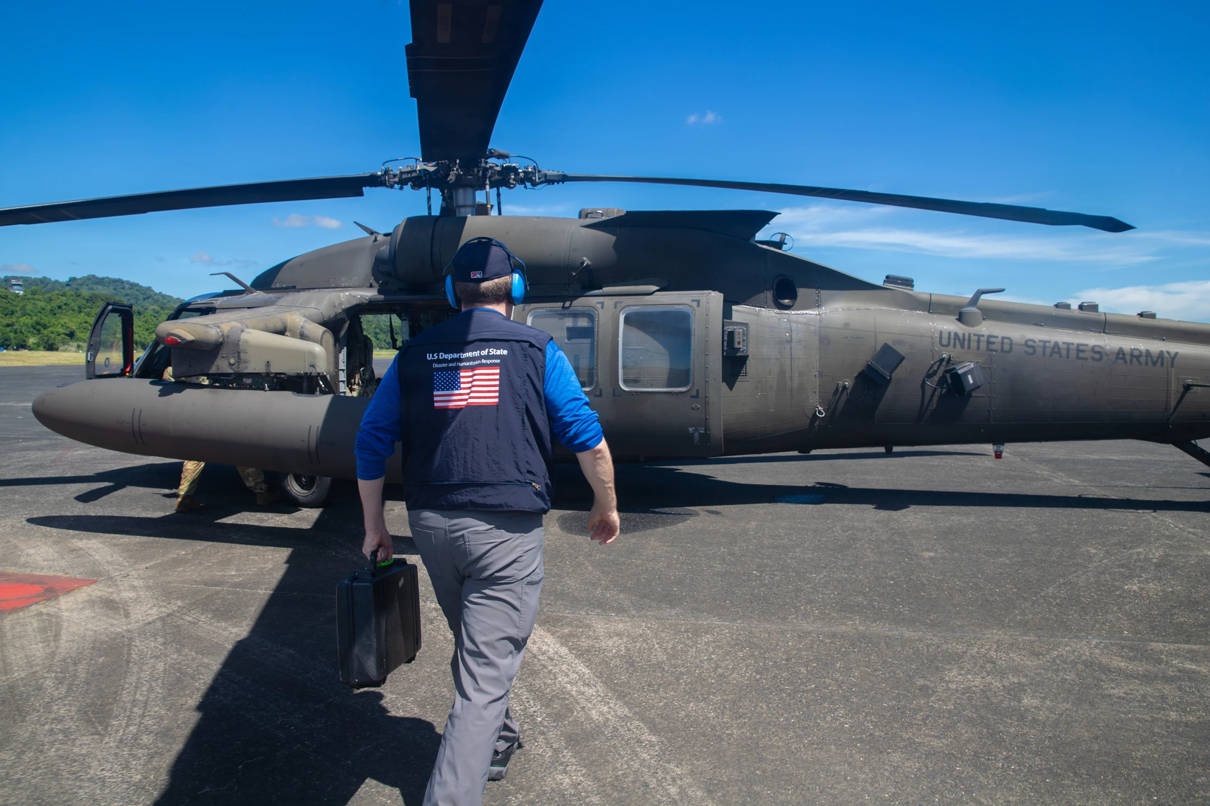 A man in a blue shirt and a vest with a U.S. flag patch walks across an airfield toward a parked UH-60 Black Hawk helicopter under a blue sky.
