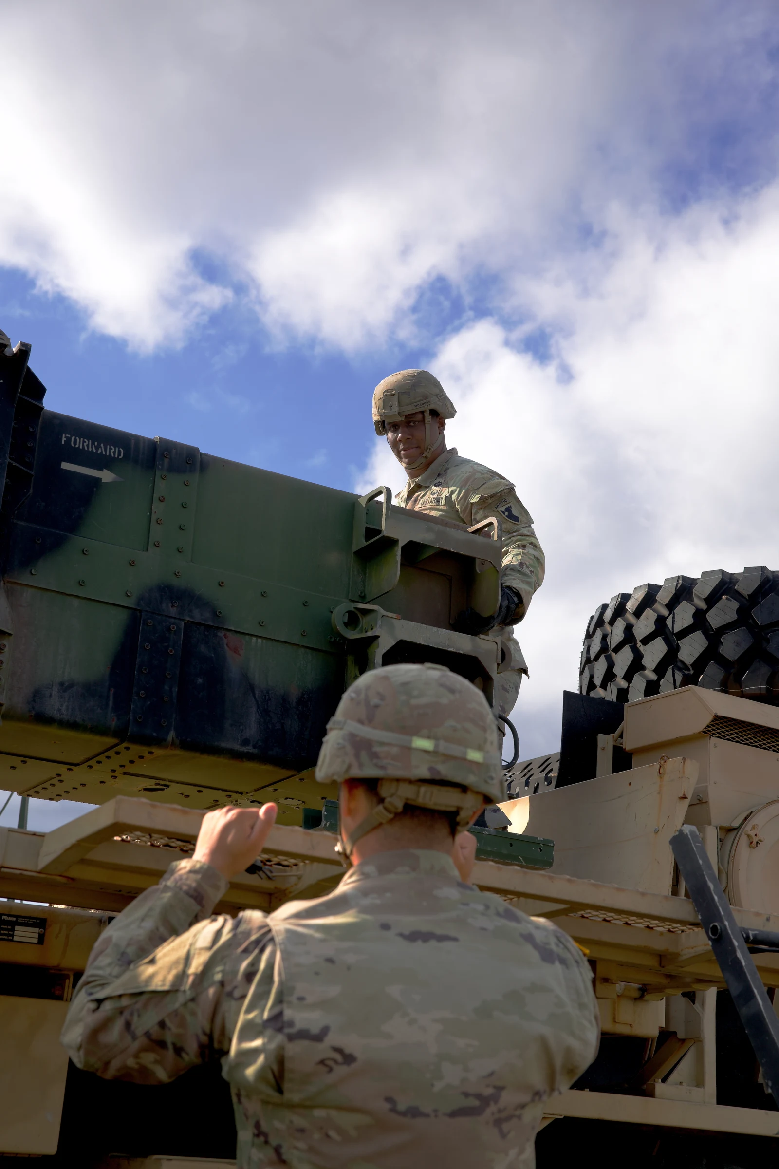 Vertical photo: two soldiers in helmets and camouflage guide a long missile canister into the frame of a Patriot launcher on a truck, one reaching up from below and one standing on top, under a blue sky with white clouds.