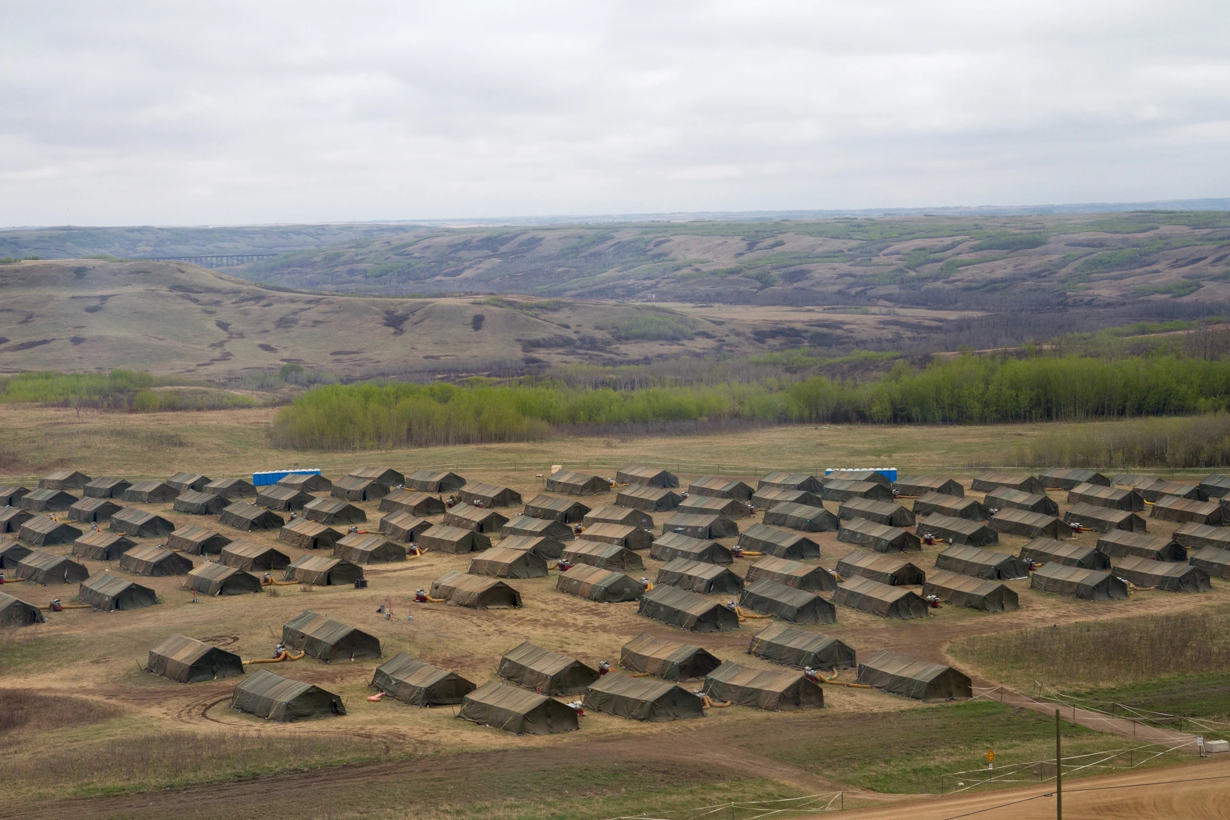Rows of olive-green military tents laid out in a field camp, seen from higher ground, with a line of trees, rolling hills and a cloudy sky beyond.