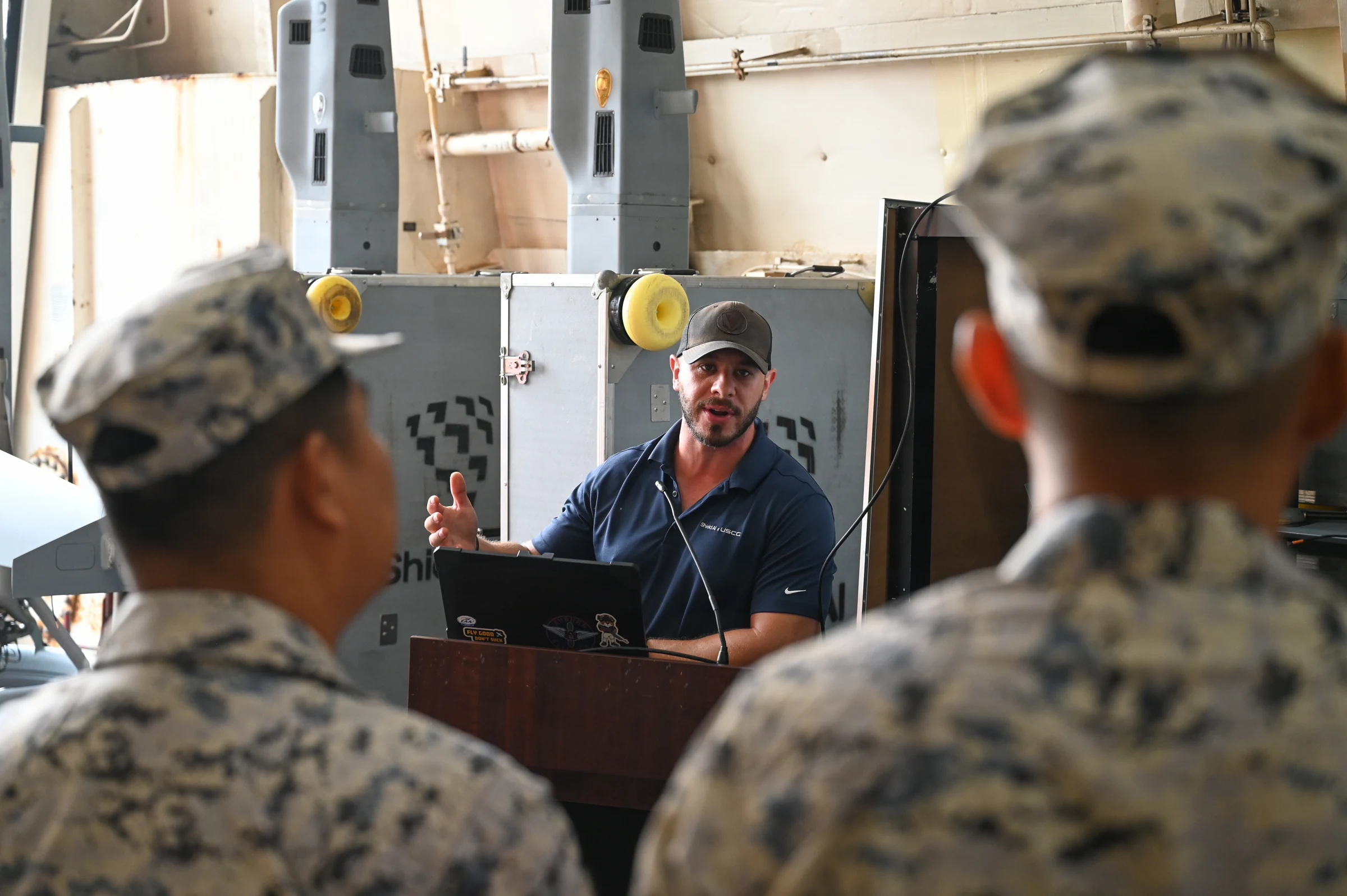 A man in a dark polo shirt and cap speaks at a lectern with a laptop inside a ship's hangar, facing two listeners in grey camouflage uniforms seen from behind; tall grey upright units stand behind him.