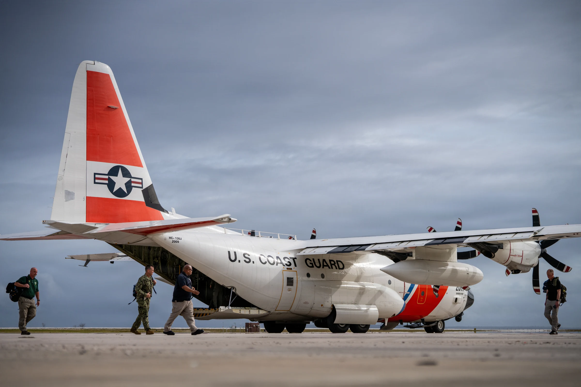 A white U.S. Coast Guard HC-130 transport plane with an orange tail stripe parked on an airport apron under grey clouds, as several people with backpacks walk past it.