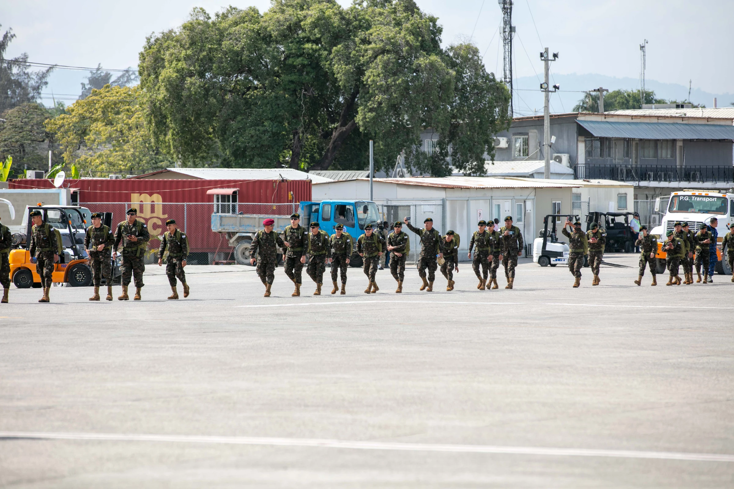 A column of about 20 soldiers in green camouflage, some in maroon or green berets, walks across a paved airport apron past parked trucks, shipping containers and low buildings, with trees and hills behind.