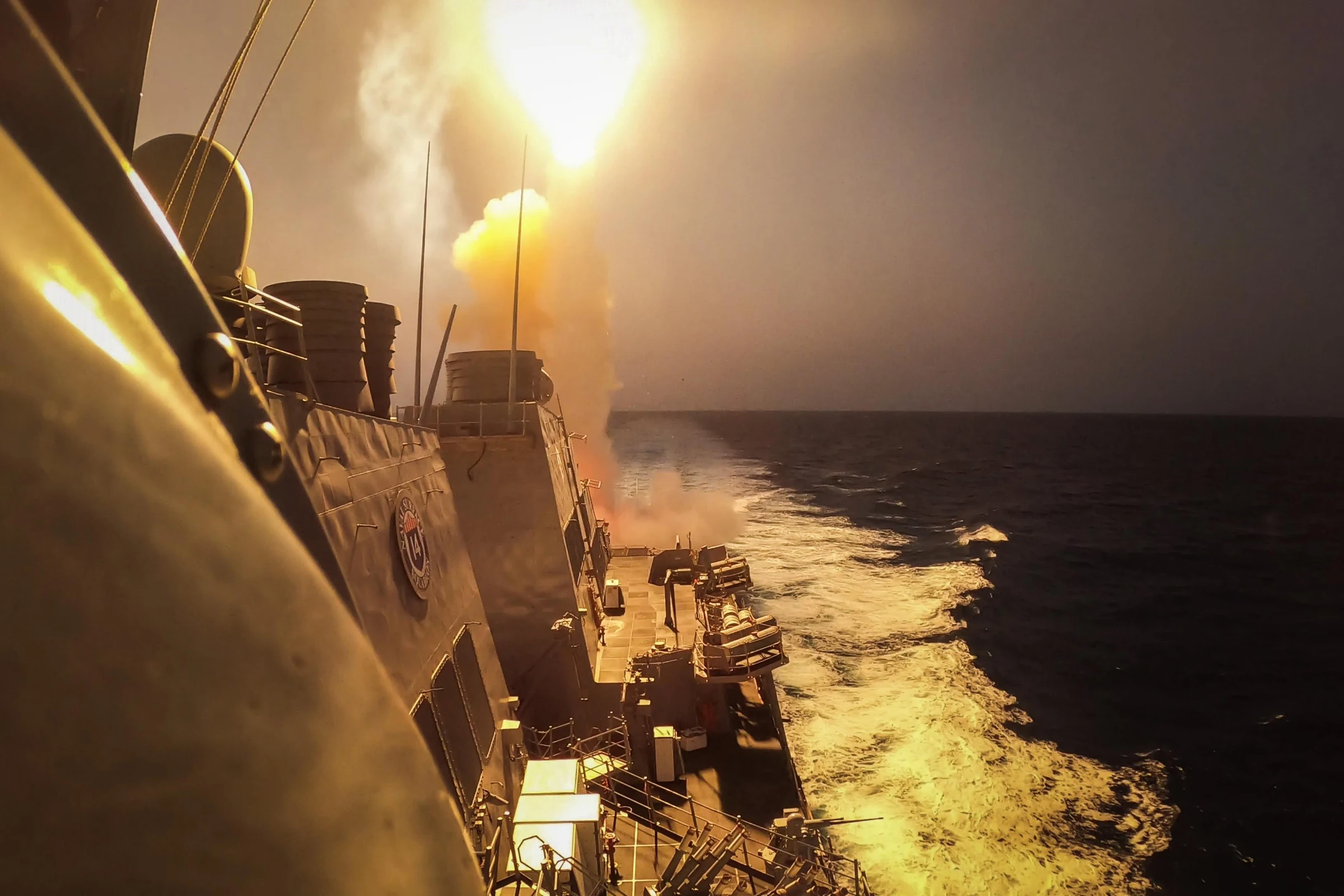 A missile launches in a bright plume from the deck of the U.S. destroyer Carney at sea, seen from along the ship's side.
