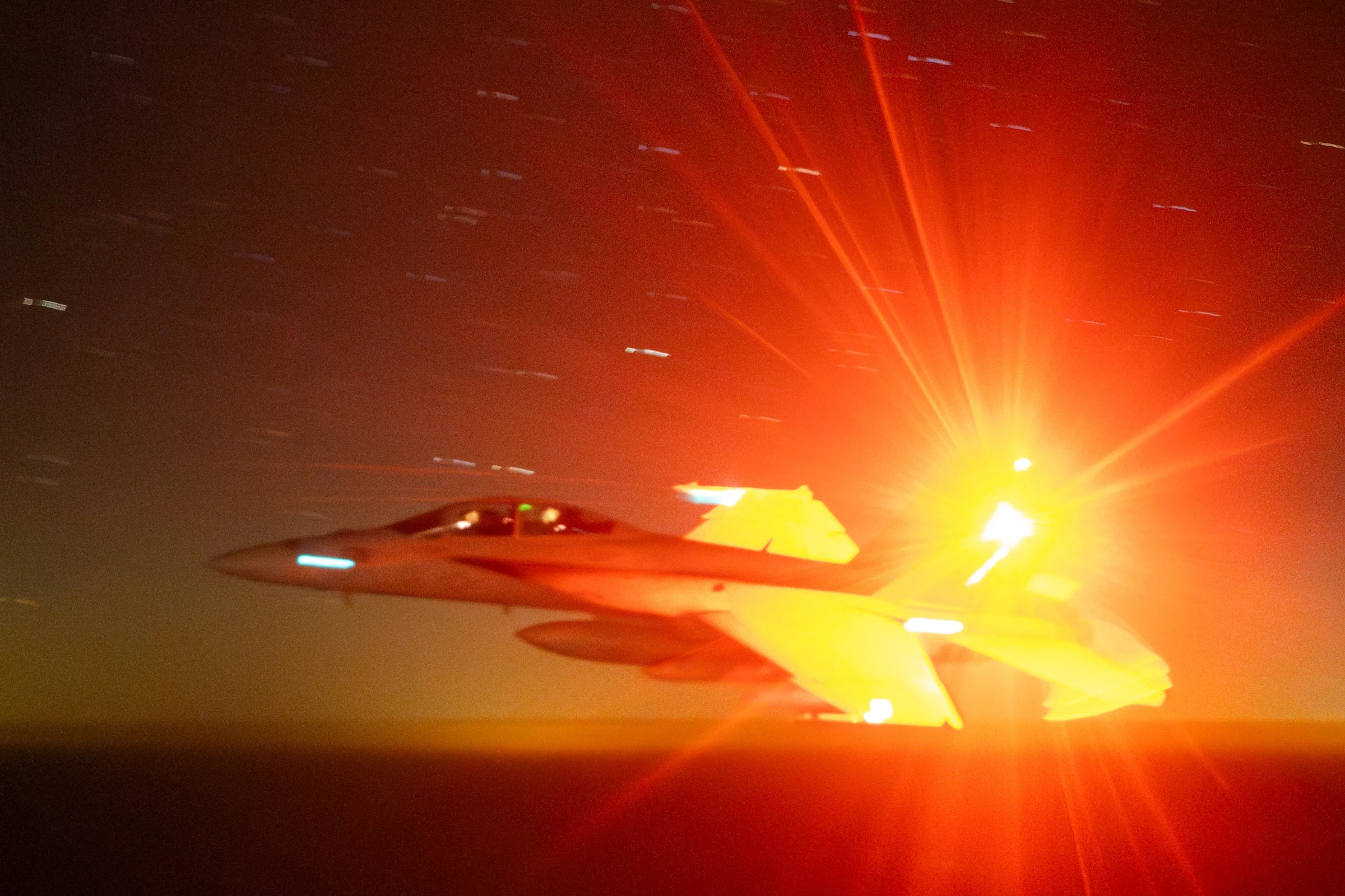 A gray U.S. Navy F/A-18 Super Hornet banks in clear sky with under-wing stores, photographed from another aircraft.