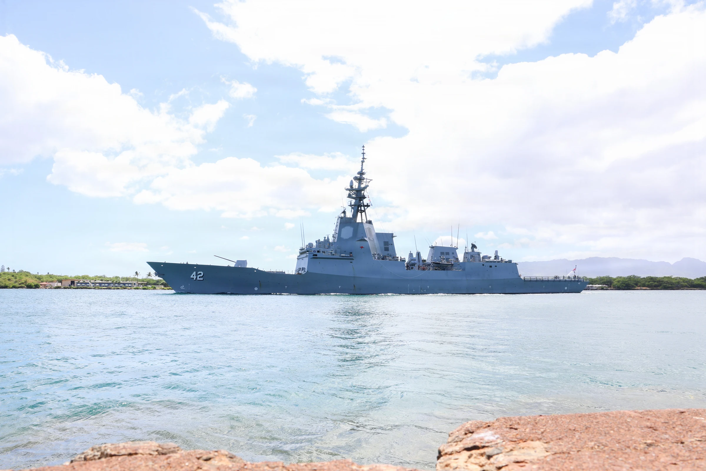 Grey Australian destroyer with hull number 42 and a tall mast sailing left to right across clear turquoise water at the Pearl Harbor entrance under white clouds, with low green shoreline and hills behind and rocks in the foreground.