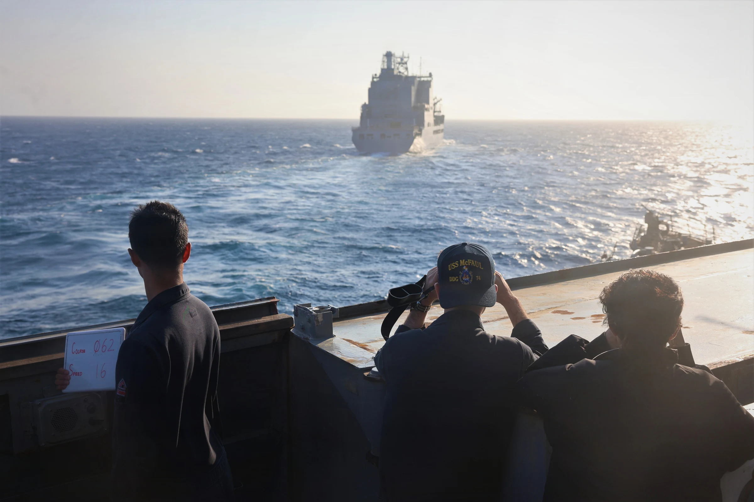 Three sailors seen from behind on a warship's bridge wing, one holding a whiteboard with course and speed numbers and one looking through binoculars, watch a large grey supply ship ahead on a sunlit sea.