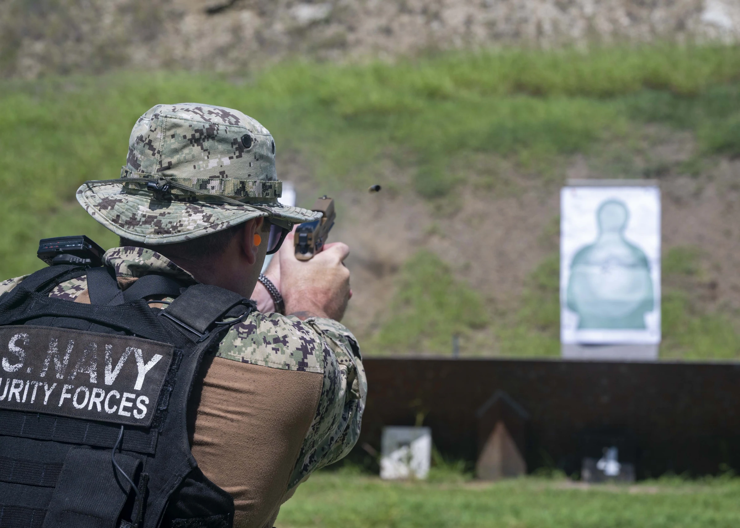 A U.S. Navy sailor in a camouflage boonie hat and a black vest reading U.S. Navy Security Forces, seen from behind, fires a pistol at a paper silhouette target on a grassy range with a dirt berm behind it.