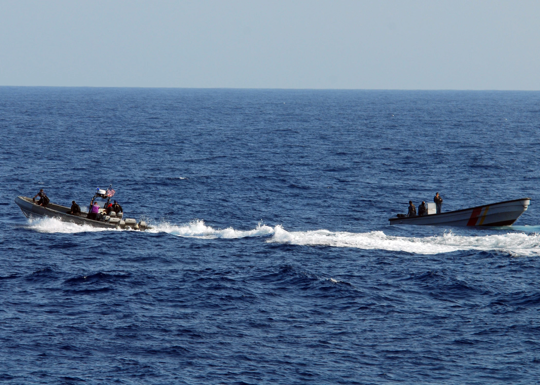 Open blue sea under a pale sky: at left a grey rigid-hull boat flying a U.S. flag carries several crew in dark gear, and at right a white Colombian navy patrol boat with red and yellow stripes carries three crew, a white wake running between them.