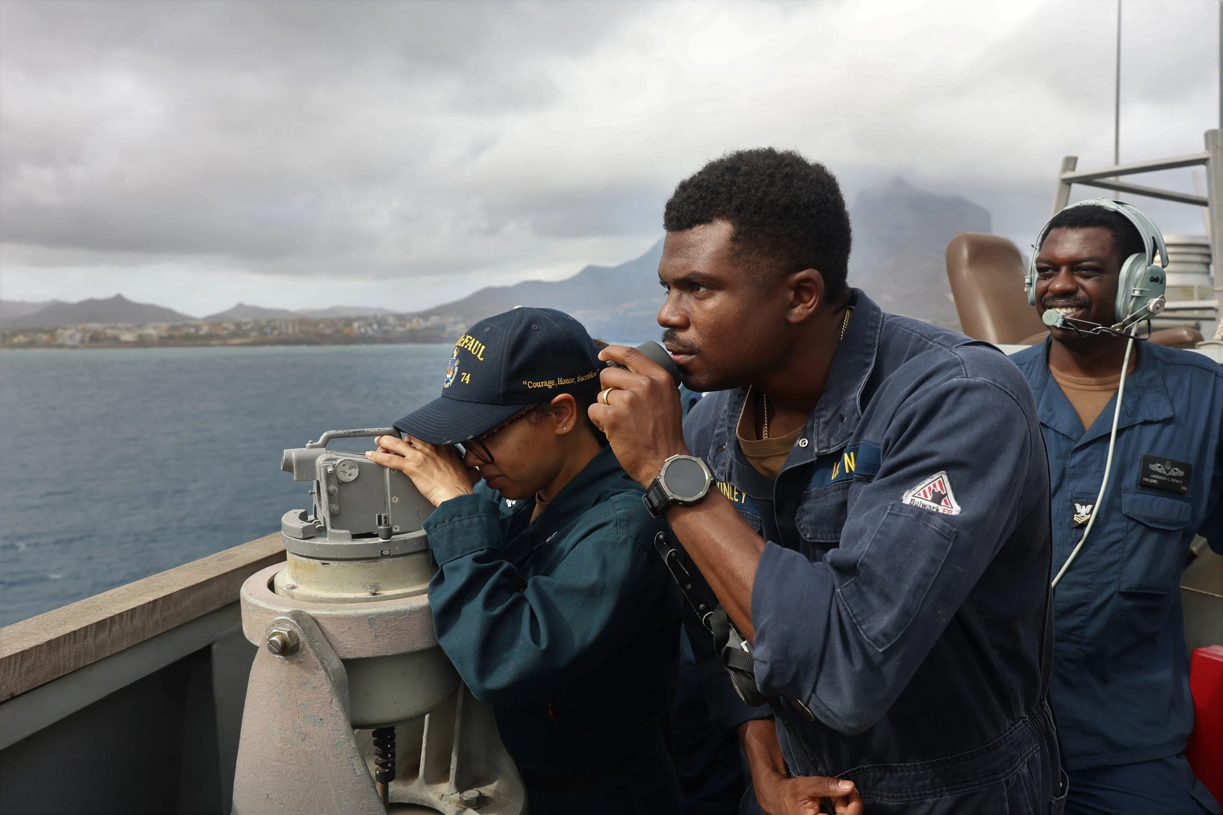 Two sailors in dark Navy uniforms look out from a warship's bridge wing, one through mounted binoculars, with a hazy harbor and hills behind them; a third sailor wearing a headset stands at right.