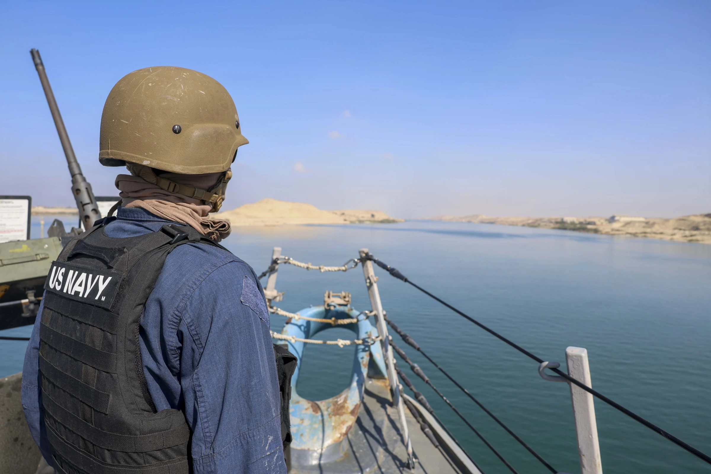 A U.S. Navy sailor in a helmet and a vest marked U.S. Navy stands at the rail of a warship's forecastle, looking out over the calm water of the Suez Canal with sandy banks behind; a mounted gun barrel rises at the left of the frame.