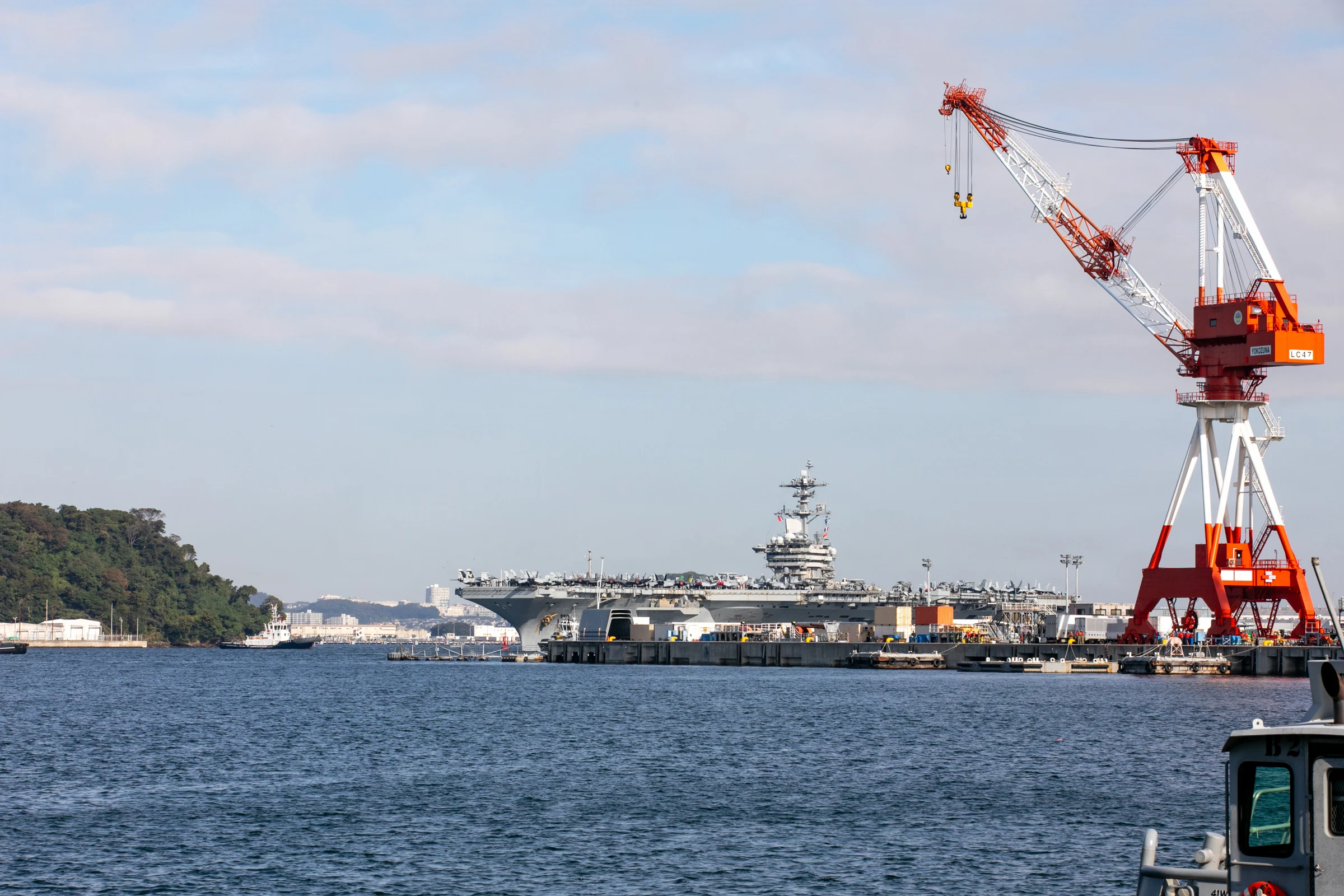 The aircraft carrier USS Theodore Roosevelt, aircraft on its deck, berthed across the harbour at Yokosuka beside a tall red-and-white crane under a pale sky.