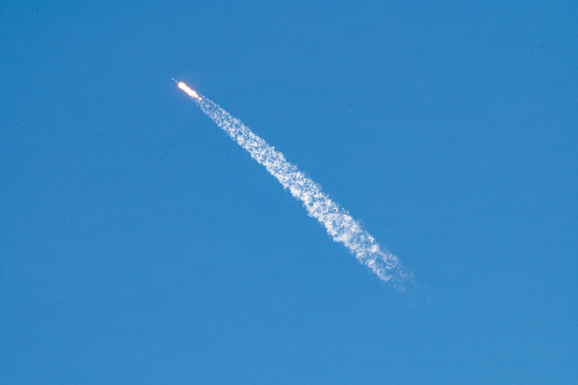 A Falcon 9 rocket climbs diagonally across a clear blue sky, its bright engine plume trailing a long white exhaust cloud.