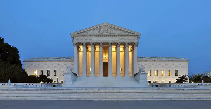 West front of the United States Supreme Court building in Washington at dusk, with columns, steps, and the flag.